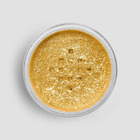A small, round container of Jenna Rae Cakes FND TESTING - 24k Magic Edible Glitter (Copy) filled with loose, shimmery gold glitter is viewed from above against a plain light gray background.