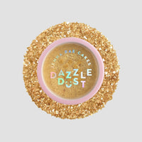 A round container of Jenna Rae Cakes FND TESTING - 24k Magic Edible Glitter (Copy), filled with gold edible glitter, is surrounded by loose gold glitter on a light grey background. The pink lid features colorful holographic text.