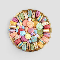 The Jenna Rae Cakes Assorted Party Platter features a round gold tray with colorful macarons, meringues, cookies, and biscuits artfully arranged in a circle on a light gray background.