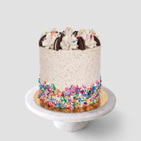 The Birthday Oreo Cake by Jenna Rae Cakes features cookies and cream frosting, whipped cream swirls, chocolate sandwich cookies on top, and colorful sprinkles around the base, beautifully displayed on a marble cake stand.