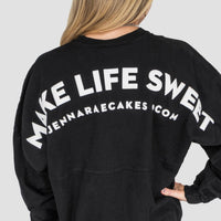 A person with long blonde hair, seen from behind, wears the Jenna Rae Cakes Black + White Spirit Jersey featuring MAKE LIFE SWEET and jennaraecakes.com in bold white letters on the back.
