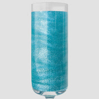 A champagne glass glows with a metallic blue shimmer from Jenna Rae Cakes Blue Sky Edible Glitter (5g Shaker, Pack of 6), set against a plain light gray background.