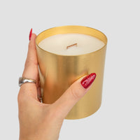 A hand with red glittery nails and a silver ring holds the Jenna Rae Cakes Brown Butter Icing Candle, featuring a metallic gold finish and wooden wick, against a plain gray background.