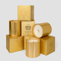 A display of Jenna Rae Cakes Brown Butter Icing Candles, featuring gold tones in metallic containers and gold-printed boxes, is arranged artistically against a plain light gray background.
