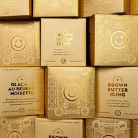Several stacked golden boxes labeled Brown Butter Icing Candle from Jenna Rae Cakes, featuring a shiny finish, decorative patterns, a smiley face, and the JR logo.