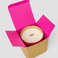 The Jenna Rae Cakes Brown Butter Icing Candle, a large white candle with a wooden wick in a gold container, is displayed in an open gold box with bright pink flaps on a light gray background.