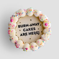 A round Jenna Rae Cakes Burn Away Cake with white frosting, colorful sprinkles, and pink accents. The center displays BURN-AWAY CAKES ARE HERE! in black icing on a smooth white background.