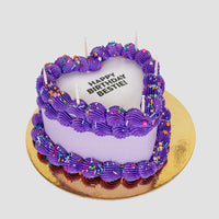 The Burn Away Heart Cake by Jenna Rae Cakes features a heart shape, purple frosting, colorful sprinkles, five unlit candles, and HAPPY BIRTHDAY BESTIE! on top. Its set on a gold cake board against a plain backdrop.