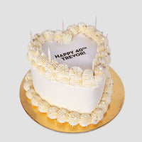 The Burn Away Heart Cake by Jenna Rae Cakes is a heart-shaped white cake with piped cream, pearl-like accents, and five unlit candles. HAPPY 40TH TREVOR! is written on top, displayed on a round gold board against a plain background.