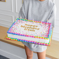 A person in a gray sweatshirt holds a Jenna Rae Cakes Classic Sheet Cake decorated with colorful icing, sprinkles, candles, and happy birthday Brian written on top.