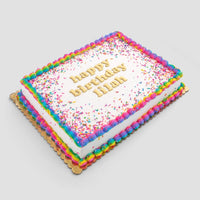 The Classic Sheet Cake by Jenna Rae Cakes features rainbow icing borders, rainbow sprinkles, and happy birthday Hilah in yellow on top. Presented on a gold board with a plain light background.