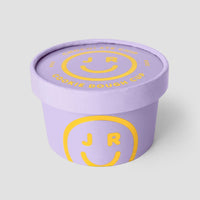 A lavender Jenna Rae Cakes Cookie Dough Cup with a matching lid, both featuring COOKIE DOUGH CUP in yellow and a smiley face with J R inside on the side and top.