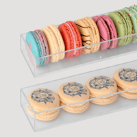 Two clear trays from Jenna Rae Cakes’ Dessert Tables feature colorful macarons in assorted flavors on top and beige macarons with intricate printed designs below, set against a plain light gray background.