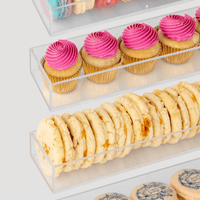 Jenna Rae Cakes Dessert Tables feature clear acrylic display shelves stocked with colorful macarons, bright pink cupcakes, crumbly cookies, and beige macarons with blue prints on a simple light background.