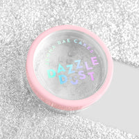 A small round container with a pink rim labeled Jenna Rae Cakes Dazzle Dust Best Seller Trio sits on a glittery silver surface, with its transparent lid revealing the shimmery powder inside.