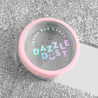 A small round container with a pink lid labeled “Jenna Rae Cakes Disco Ball Edible Glitter - 5g Shaker - Package of 6” sits on a sparkling silver glitter surface.