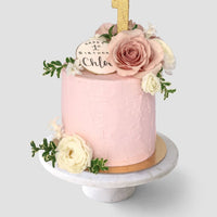 The Fresh Floral Buttercream Cake by Jenna Rae Cakes features pink frosting, fresh flowers, a gold glitter number 1 topper, and a round Happy 1st Birthday Chloe plaque, beautifully presented on a white cake stand.