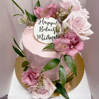 The Fresh Floral Buttercream Cake by Jenna Rae Cakes features pink frosting, fresh pink roses, green leaves, and a white Happy Bday Milana topper in black script, displayed on a gold cake board.