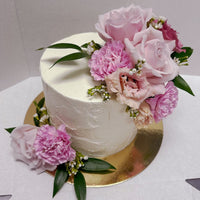 The Fresh Floral Buttercream Cake by Jenna Rae Cakes features white frosting, pink roses, carnations, small white flowers, and green leaves on a gold cake board.