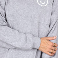 A person wears the Grey + White Spirit Jersey by Jenna Rae Cakes, featuring a white smiley face and the letters J and R on the chest, with their arms crossed and rings visible on their fingers.