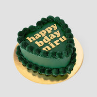 The Heart Cake by Jenna Rae Cakes features a dark green, heart-shaped design with wavy piped borders and “happy bday niru” in yellow icing, set on a gold board against a light background.