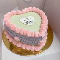 A heart-shaped cake with pink and purple icing borders on a gold base. A white heart plaque on top says Happy Birthday, Love in black script. The cake is inside an open white box.