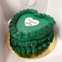 A heart-shaped cake with green frosting and decorative swirls, displayed on a gold cake board. A white heart-shaped plaque on top reads happy year in cursive, all inside a white cake box.