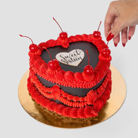 The Heart Cake by Jenna Rae Cakes features red frosting, a black icing center with Sweet Sixteen, topped with cherries. A hand places a cherry on top as the cake sits on a gold board against a light background.