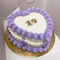 A heart-shaped cake with purple and white frosting, decorated with the number 19 in gold on top, placed on a gold cake board inside a white box.