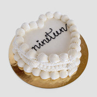 The Heart Cake by Jenna Rae Cakes is a white, heart-shaped cake with decorative icing borders and nineteen written in black icing, set on a round gold cake board.