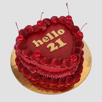 The Heart Cake by Jenna Rae Cakes is a heart-shaped red cake with decorative red frosting, six cherries on top, and “hello 21” in gold icing, presented on a round golden board.