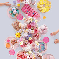 A festive dessert table inspired by Jenna Rae Cakes at Home Cookbook displays pink plates, colorful pastries, waffles, and macarons as children reach for treats. The bright scene captures the playful spirit of Jenna Rae Cakes.