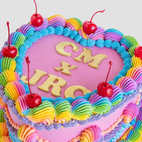 A pink heart-shaped cake from Jenna Rae Cakes, topped with rainbow frosting, red cherries, and yellow letters spelling CM x JRC using the Jenna Rae Cakes x Colour Mill Pack.