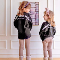 Two young girls smile at each other in front of a white wall and macaron photo, both wearing matching Jenna Rae Cakes Kids Black Spirit Jersey shirts, tan leggings, boots, and headbands.