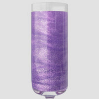 A clear champagne flute contains sparkling, shimmery purple liquid made with Jenna Rae Cakes Lavender Haze Edible Glitter (5g Shaker, Package of 6) against a plain light gray background.