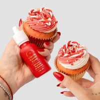 Two hands with red nails hold cupcakes with red and white swirled frosting and sprinkles. One hand also holds a small “Liquid Candy Cane - Package of 6” bottle by Jenna Rae Cakes. The background is light gray.