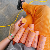 Wearing bright orange pleated pants, a person holds a round container of Jenna Rae Cakes Liquid Candy Peach and has several matching tubes on their lap, accessorized with bracelets and black sandals beside a yellow cord.