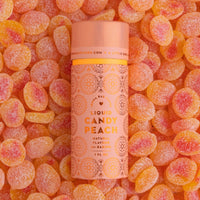 A cylindrical Liquid Candy Peach container from Jenna Rae Cakes rests on orange and yellow peach-shaped gummy candies, featuring a peach-colored design with floral patterns.