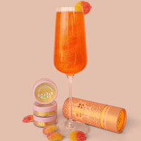 A Jenna Rae Cakes Liquid Candy Peach sparkling orange drink is served in a champagne flute with gummy candies inside and on the rim, next to “Dazzle Dust” containers and a cylindrical package, all set on a peach-colored background.