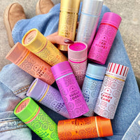 A person in jeans holds a package of six colorful Liquid Golden Bubbly bottles from Jenna Rae Cakes, featuring dessert-inspired scents. Brown shoes appear in the background.