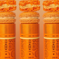 Three orange Jenna Rae Cakes Liquid Pumpkin Spice canisters are each topped with a pumpkin macaron and pumpkin-flavored cookie, featuring decorative patterns and branding on a light background.