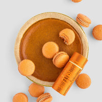A pumpkin pie surrounded by orange macarons, one partially broken, is shown with a Jenna Rae Cakes Liquid Pumpkin Spice container on a light gray background.