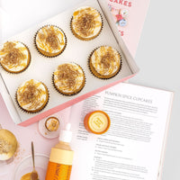 A package of six Jenna Rae Cakes Liquid Pumpkin Spice sits next to an open cookbook with a pumpkin spice cupcake recipe, along with a frosting bottle, small jar, spoon, and colorful sprinkles scattered on the table.