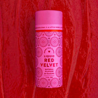 A pink bottle of Jenna Rae Cakes Liquid Red Velvet stands upright against a textured red background with swirls. The packaging features circular decorative designs and bold red lettering.