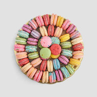 The Jenna Rae Cakes Macaron Party Platter features colorful macarons arranged in concentric circles around round cookies at the center, all beautifully displayed on a white background.