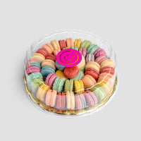 The Jenna Rae Cakes Macaron Party Platter features colorful macarons arranged in circles inside a round clear plastic container, topped with a pink smiley face decoration, set against a light gray background.