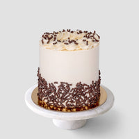 The Marble Cake by Jenna Rae Cakes is a tall, round cake with smooth white frosting, topped with icing swirls and chocolate shavings around the base, displayed on a white marble cake stand against a plain background.
