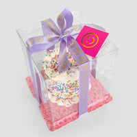 The Mini Vanilla Sprinkle Cake by Jenna Rae Cakes features white frosting with colorful sprinkles in a clear box, accented with a lavender ribbon of yellow smiley faces, pink smiley-face tag, and a pink patterned base.