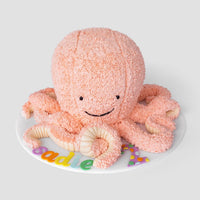 A pink, octopus-shaped cake with textured frosting, a smiling face, and curled tentacles sits on a white round board decorated with colorful letters.