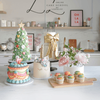 A vibrant kitchen display showcases Jenna Rae Cakes Online Classes with cakes like a unicorn cake, pink floral cake, Christmas tree cake, and pastel cupcakes on a marble counter, accented by flowers and contemporary decor.